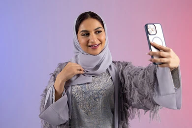 الاحتفاظ باللحظات السعيدة، التقاط صورة سيلفي تذكارية عبر جهاز الجوال بإيماءات السعادة والسرور، بورتريه لامرأة عربية خليجية اماراتية محجبة ترتدي العباءة تمسك بيدها الهاتف المحمول، شابة سعودية تستخدم جهاز تقني حديث ومتطور، خلفية ملونة
