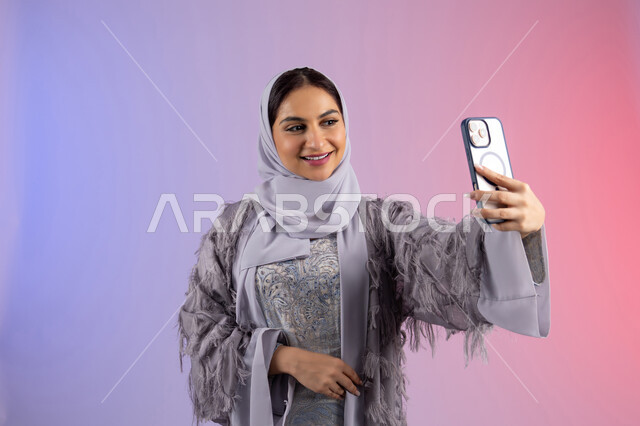 Preserving happy moments, taking a commemorative selfie with a mobile phone with gestures of happiness and pleasure, portrait of a veiled Emirati Gulf Arab woman wearing an abaya holding a mobile phone, a young Saudi woman using a modern and advanced technical device, colorful background