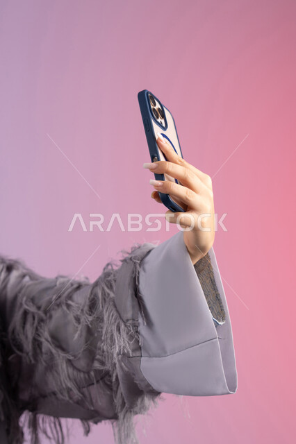 شابة سعودية تستخدم جهاز تقني حديث ومتطور، الاحتفاظ باللحظات السعيدة، التقاط صورة سيلفي تذكارية عبر جهاز الجوال، بورتريه مقرب ليد لامرأة عربية خليجية اماراتية ترتدي العباءة تمسك بيدها الهاتف المحمول، خلفية ملونة