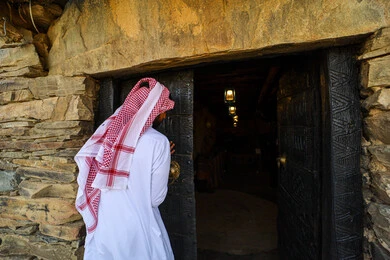 Old-style architectural art of museums, historical forts and castles, stone buildings and structures, famous cultural heritage sites and landmarks, a back view of a Saudi Arabian Gulf man wearing a traditional shemagh and thobe walking around the Lod Heritage Museum in Al-Baha region
