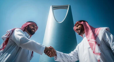 Greetings, peace and agreement on a successful business deal, two Saudi Arabian Gulf businessmen wearing traditional thobe and shemagh standing outside the company headquarters shaking hands with expressions of joy and happiness, Kingdom Tower background