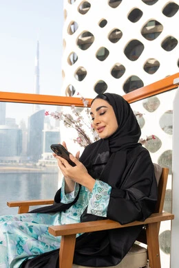 تصفح التطبيقات وبرامج التواصل الاجتماعي، تبادل الأحاديث والأخبار مع الأهل والأصدقاء،  امراة عربية خليجية اماراتية ترتدي العباءة والحجاب تمسك بيدها الهاتف المحمول تجلس في الخارج، استخدام جهاز تقني حديث ومتطور