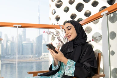 Expressions of happiness and integration, browsing social media applications and programs, exchanging conversations and news with family and friends, an Arab Gulf Emirati woman wearing an abaya and hijab holding a mobile phone in her hand, sitting outside, using a modern and advanced technical device