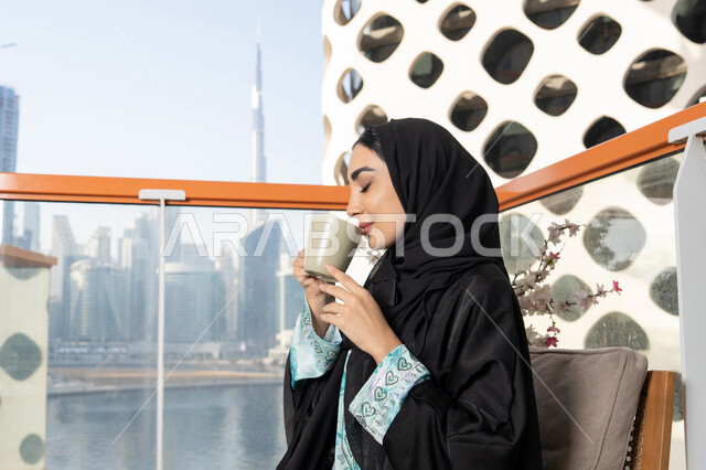 تعابير المتعة والاسترخاء، قضاء وقت الفراغ باحتساء المشروب المفضل, تناول الأعشاب الشتوية الدافئة، امراة عربية خليجية اماراتية ترتدي العباءة والحجاب تقوم بشرب القهوة، الاستمتاع بالمشروبات الساخنة