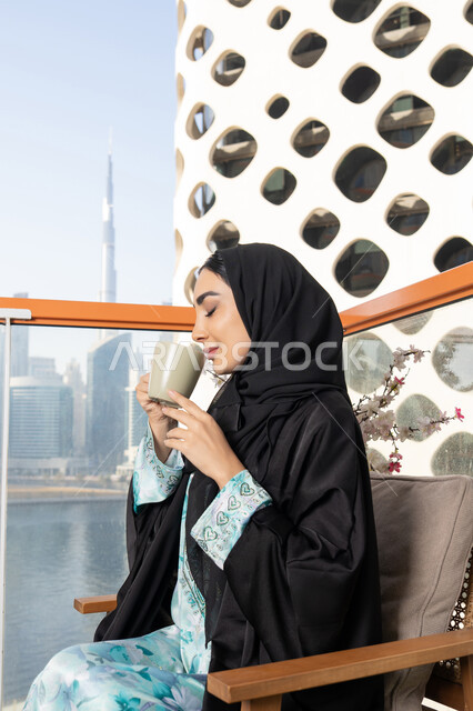Enjoying hot drinks, gestures of pleasure and relaxation, spending free time sipping a favorite drink, eating warm winter herbs, an Arab Gulf Emirati woman wearing an abaya and hijab drinking coffee