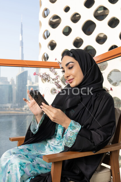 تعابير السرور والاندماج، تصفح التطبيقات وبرامج التواصل الاجتماعي، تبادل الأحاديث والأخبار مع الأهل والأصدقاء،  امراة عربية خليجية اماراتية ترتدي العباءة والحجاب تمسك بيدها الهاتف المحمول تجلس في الخارج، استخدام جهاز تقني حديث ومتطور