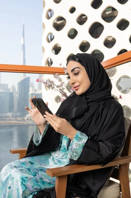 تعابير السرور والاندماج، تصفح التطبيقات وبرامج التواصل الاجتماعي، تبادل الأحاديث والأخبار مع الأهل والأصدقاء،  امراة عربية خليجية اماراتية ترتدي العباءة والحجاب تمسك بيدها الهاتف المحمول تجلس في الخارج، استخدام جهاز تقني حديث ومتطور