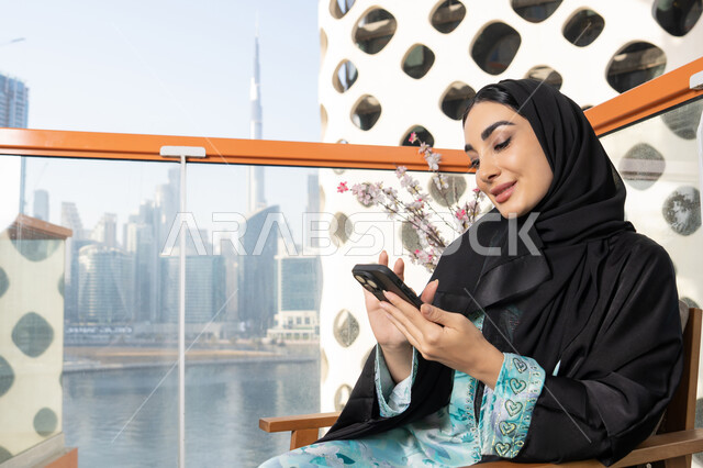 Browsing social media applications and programs, exchanging conversations and news with family and friends, an Emirati Gulf Arab woman wearing an abaya and hijab holding a mobile phone in her hand, sitting outside, using a modern and advanced technical device