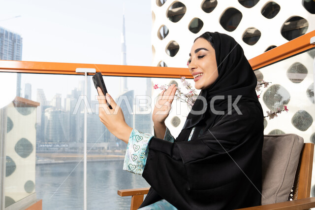 ايماءات السعادة والسرور، اجراء مكالمة فيديو عبر جهاز الجوال، التواصل وتبادل الأحاديث والأخبار مع الأهل والأصدقاء، امراة عربية خليجية اماراتية ترتدي العباءة والحجاب تمسك بيدها الهاتف المحمول، استخدام جهاز تقني حديث ومتطور، قضاء وقت ممتع في الخارج