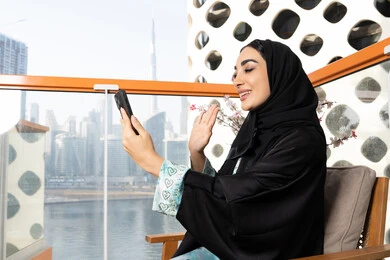 ايماءات السعادة والسرور، اجراء مكالمة فيديو عبر جهاز الجوال، التواصل وتبادل الأحاديث والأخبار مع الأهل والأصدقاء، امراة عربية خليجية اماراتية ترتدي العباءة والحجاب تمسك بيدها الهاتف المحمول، استخدام جهاز تقني حديث ومتطور، قضاء وقت ممتع في الخارج