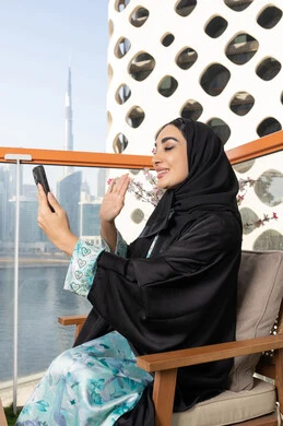 Having a good time outside, gestures of happiness and pleasure, making a video call via mobile device, communicating and exchanging conversations and news with family and friends, an Arab Gulf Emirati woman wearing an abaya and hijab holding a mobile phone in her hand, using a modern and advanced technical device