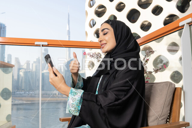 قضاء وقت ممتع في الخارج، ايماءات السعادة والسرور، اجراء مكالمة فيديو عبر جهاز الجوال، التواصل وتبادل الأحاديث والأخبار مع الأهل والأصدقاء، امراة عربية خليجية اماراتية ترتدي العباءة والحجاب تمسك بيدها الهاتف المحمول، استخدام جهاز تقني حديث ومتطور