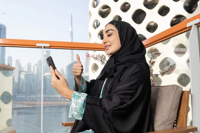 Having a good time outside, gestures of happiness and pleasure, making a video call via mobile device, communicating and exchanging conversations and news with family and friends, an Arab Gulf Emirati woman wearing an abaya and hijab holding a mobile phone in her hand, using a modern and advanced technical device
