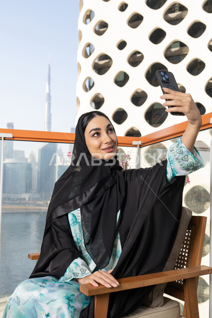 توثيق اللحظات التذكارية السعيدة، استخدام جهاز تقني حديث ومتطور لتخليد الذكريات ، التقاط الصور الشخصية باستخدام الهاتف المحمول، امراة عربية خليجية اماراتية ترتدي العباءة والحجاب تأخذ صورة سيلفي بإيماءات السعادة والسرور