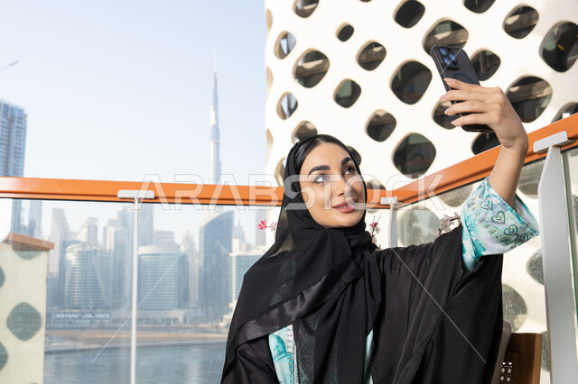 Documenting happy memorable moments, using a modern and advanced technical device to immortalize memories, taking selfies using a mobile phone, an Emirati Gulf Arab woman wearing an abaya and hijab taking a selfie with gestures of happiness and pleasure
