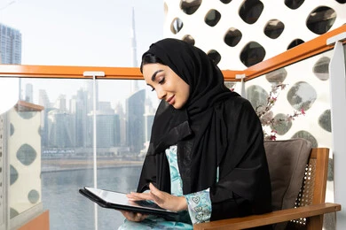 مهنة ووظيفة نسائية، إمرأة عربية خليجية اماراتية ترتدي العباءة والحجاب تستخدم جهاز التابليت المحمول، استخدام جهاز تقني حديث ومتطور، العمل عن بعد في الخارج، ادارة الأعمال وانجاز المهام المطلوبة عبر الجهاز اللوحي