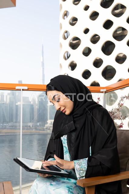 ادارة الأعمال وانجاز المهام المطلوبة عبر الجهاز اللوحي، مهنة ووظيفة نسائية، إمرأة عربية خليجية اماراتية ترتدي العباءة والحجاب تستخدم جهاز التابليت المحمول، استخدام جهاز تقني حديث ومتطور، العمل عن بعد في الخارج