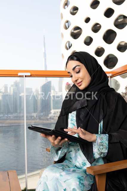 العمل عن بعد في الخارج، ادارة الأعمال وانجاز المهام المطلوبة عبر الجهاز اللوحي، مهنة ووظيفة نسائية، إمرأة عربية خليجية اماراتية ترتدي العباءة والحجاب تستخدم جهاز التابليت المحمول، استخدام جهاز تقني حديث ومتطور