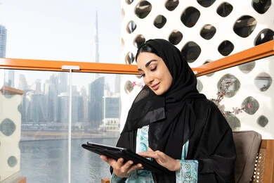 مهنة ووظيفة نسائية، إمرأة عربية خليجية اماراتية ترتدي العباءة والحجاب تستخدم جهاز التابليت المحمول، استخدام جهاز تقني حديث ومتطور، العمل عن بعد في الخارج، ادارة الأعمال وانجاز المهام المطلوبة عبر الجهاز اللوحي