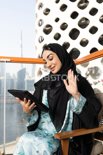 اجراء مكالمة فيديو، ادارة الأعمال وانجاز المهام المطلوبة عبر الجهاز اللوحي، مهنة ووظيفة نسائية، إمرأة عربية خليجية اماراتية ترتدي العباءة والحجاب تستخدم جهاز التابليت المحمول، استخدام جهاز تقني حديث ومتطور، العمل عن بعد في الخارج