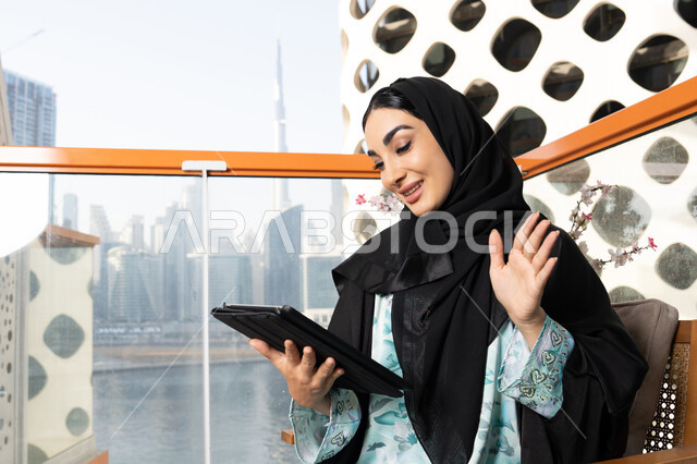 اجراء مكالمة فيديو، ادارة الأعمال وانجاز المهام المطلوبة عبر الجهاز اللوحي، مهنة ووظيفة نسائية، إمرأة عربية خليجية اماراتية ترتدي العباءة والحجاب تستخدم جهاز التابليت المحمول، استخدام جهاز تقني حديث ومتطور، العمل عن بعد في الخارج