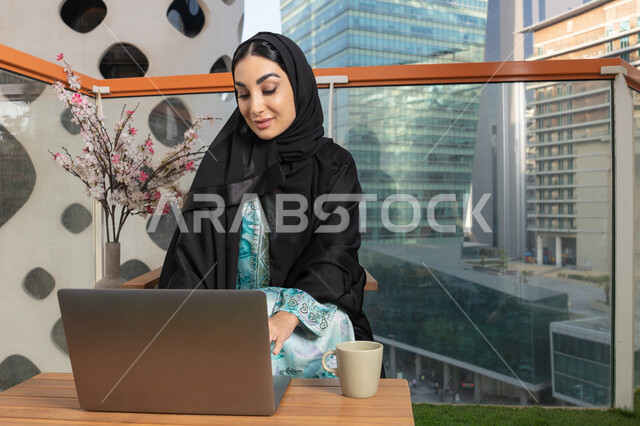 ايماءات التركيز والاندماج، ادارة الأعمال وانجاز المهام المطلوبة، العمل عن بعد،  مهنة ووظيفة نسائية، إمرأة عربية خليجية اماراتية محجبة ترتدي العباءة والحجاب تستخدم الحاسوب المحمول، استخدام جهاز تقني حديث ومتطور