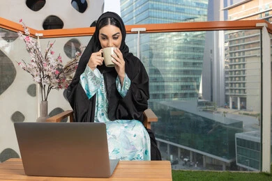 العمل على الحاسوب المحمول، الاستمتاع بالمشروبات الساخنة، تعابير المتعة والاسترخاء، قضاء وقت الفراغ باحتساء المشروب المفضل، تناول الأعشاب الشتوية الدافئة، امراة عربية خليجية اماراتية ترتدي العباءة والحجاب تقوم بشرب القهوة واستخدام جهاز تقني حديث