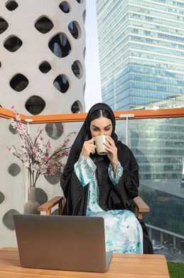 Using a modern technical device, eating warm winter herbs, an Emirati Gulf Arab woman wearing an abaya and hijab drinking coffee and working on a laptop, enjoying hot drinks, expressions of pleasure and relaxation, spending free time sipping a favorite drink