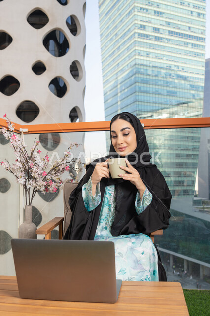 استخدام جهاز تقني حديث، تناول الأعشاب الشتوية الدافئة، امراة عربية خليجية اماراتية ترتدي العباءة والحجاب تقوم بشرب القهوة والعمل على الحاسوب المحمول، الاستمتاع بالمشروبات الساخنة، تعابير المتعة والاسترخاء، قضاء وقت الفراغ باحتساء المشروب المفضل