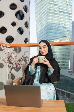 استخدام جهاز تقني حديث، تناول الأعشاب الشتوية الدافئة، امراة عربية خليجية اماراتية ترتدي العباءة والحجاب تقوم بشرب القهوة والعمل على الحاسوب المحمول، الاستمتاع بالمشروبات الساخنة، تعابير المتعة والاسترخاء، قضاء وقت الفراغ باحتساء المشروب المفضل