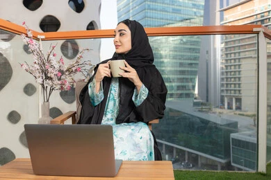 Working on laptop, enjoying hot drinks, expressions of pleasure and relaxation, spending free time sipping favorite drink, eating warm winter herbs, an Arab Gulf Emirati woman wearing abaya and hijab drinking coffee and using a modern technical device