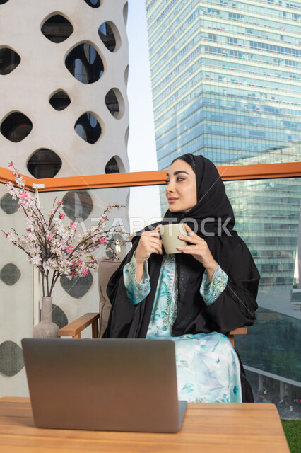 Using a modern technical device, eating warm winter herbs, an Emirati Gulf Arab woman wearing an abaya and hijab drinking coffee and working on a laptop, enjoying hot drinks, expressions of pleasure and relaxation, spending free time sipping a favorite drink
