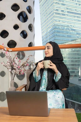 Using a modern technical device, eating warm winter herbs, an Emirati Gulf Arab woman wearing an abaya and hijab drinking coffee and working on a laptop, enjoying hot drinks, expressions of pleasure and relaxation, spending free time sipping a favorite drink