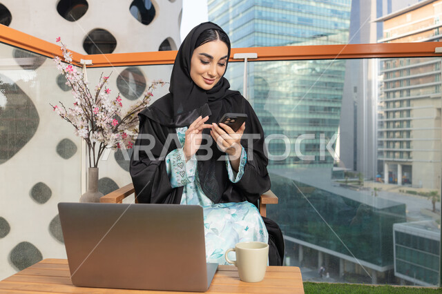 Exchanging conversations and news with family and friends, an Emirati woman wearing an abaya and hijab holding a mobile phone and drinking coffee, using a modern and advanced technical device, sipping her favorite drink in a cafe, browsing applications and social networking programs