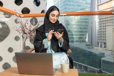 تبادل الأحاديث والأخبار مع الأهل والأصدقاء،   امراة اماراتية ترتدي العباءة والحجاب تمسك بيدها الهاتف المحمول وتشرب القهوة، استخدام جهاز تقني حديث ومتطور، احتساء المشروب المفضل في المقهى، تصفح التطبيقات وبرامج التواصل الاجتماعي