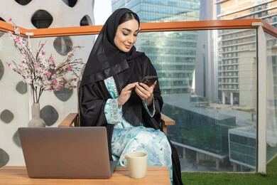 تصفح التطبيقات وبرامج التواصل الاجتماعي، تبادل الأحاديث والأخبار مع الأهل والأصدقاء،   امراة اماراتية ترتدي العباءة والحجاب تمسك بيدها الهاتف المحمول وتشرب القهوة، استخدام جهاز تقني حديث ومتطور، احتساء المشروب المفضل في المقهى