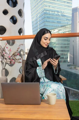 Exchanging conversations and news with family and friends, an Emirati woman wearing an abaya and hijab holding a mobile phone and drinking coffee, using a modern and advanced technical device, sipping her favorite drink in a cafe, browsing applications and social networking programs