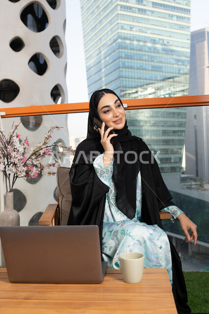 Making a phone call on a mobile device, communicating and exchanging conversations and news with family and friends, an Arab Gulf Emirati woman wearing an abaya and hijab holding a mobile phone in her hand and drinking coffee, using a modern and advanced technical device, sipping her favorite drink in a cafe