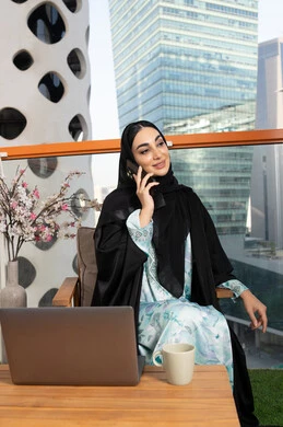Making a phone call on a mobile device, communicating and exchanging conversations and news with family and friends, an Arab Gulf Emirati woman wearing an abaya and hijab holding a mobile phone in her hand and drinking coffee, using a modern and advanced technical device, sipping her favorite drink in a cafe