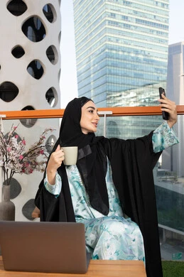 العمل على الحاسب المحمول، امراة عربية خليجية اماراتية ترتدي العباءة والحجاب تمسك بيدها الهاتف المحمول وتشرب القهوة، استخدام جهاز تقني حديث ومتطور، احتساء المشروب المفضل في المقهى، اجراء مكالمة فيديو عبر جهاز الجوال