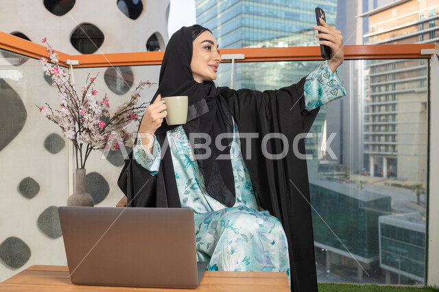 اجراء مكالمة فيديو عبر جهاز الجوال، العمل على الحاسب المحمول، امراة عربية خليجية اماراتية ترتدي العباءة والحجاب تمسك بيدها الهاتف المحمول وتشرب القهوة، استخدام جهاز تقني حديث ومتطور، احتساء المشروب المفضل في المقهى