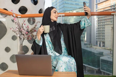 Making a video call via mobile device, working on a laptop, an Arab Gulf Emirati woman wearing an abaya and hijab holding a mobile phone and drinking coffee, using a modern and advanced technical device, sipping her favorite drink in a cafe