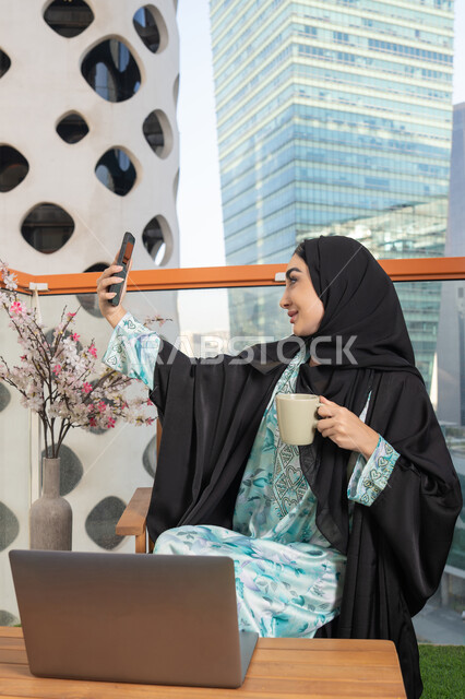 استخدام جهاز تقني حديث ومتطور، احتساء المشروب المفضل في المقهى، اجراء مكالمة فيديو عبر جهاز الجوال، العمل على الحاسب المحمول، امراة عربية خليجية اماراتية ترتدي العباءة والحجاب تمسك بيدها الهاتف المحمول وتشرب القهوة