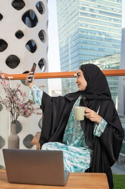 استخدام جهاز تقني حديث ومتطور، احتساء المشروب المفضل في المقهى، اجراء مكالمة فيديو عبر جهاز الجوال، العمل على الحاسب المحمول، امراة عربية خليجية اماراتية ترتدي العباءة والحجاب تمسك بيدها الهاتف المحمول وتشرب القهوة