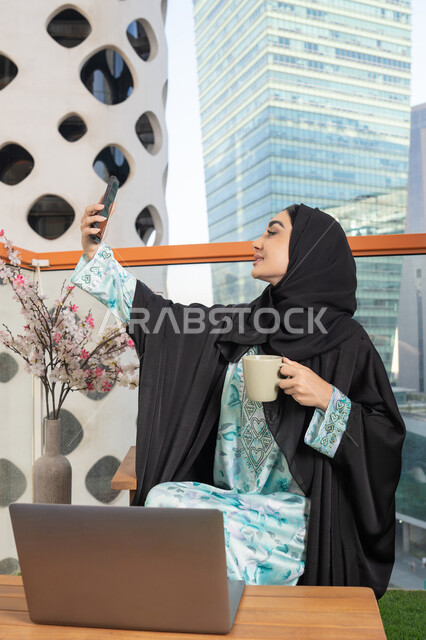 استخدام جهاز تقني حديث ومتطور، احتساء المشروب المفضل في المقهى، اجراء مكالمة فيديو عبر جهاز الجوال، العمل على الحاسب المحمول، امراة عربية خليجية اماراتية ترتدي العباءة والحجاب تمسك بيدها الهاتف المحمول وتشرب القهوة
