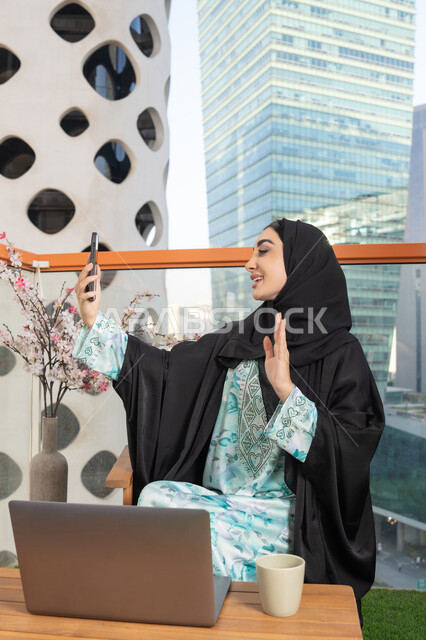 العمل على الحاسب المحمول، امراة عربية خليجية اماراتية ترتدي العباءة والحجاب تمسك بيدها الهاتف المحمول وتشرب القهوة، استخدام جهاز تقني حديث ومتطور، احتساء المشروب المفضل في المقهى، اجراء مكالمة فيديو عبر جهاز الجوال