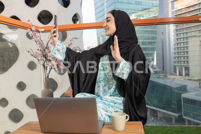 اجراء مكالمة فيديو عبر جهاز الجوال، العمل على الحاسب المحمول، امراة عربية خليجية اماراتية ترتدي العباءة والحجاب تمسك بيدها الهاتف المحمول وتشرب القهوة، استخدام جهاز تقني حديث ومتطور، احتساء المشروب المفضل في المقهى