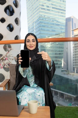 الاشارة الى شاشة سوداء فارغة عبر جهاز الجوال، العمل على الحاسب المحمول، امراة عربية خليجية اماراتية ترتدي العباءة والحجاب تمسك بيدها الهاتف المحمول وتشرب القهوة، استخدام جهاز تقني حديث ومتطور، احتساء المشروب المفضل في المقهى