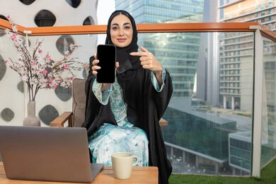 الاشارة الى شاشة سوداء فارغة عبر جهاز الجوال، العمل على الحاسب المحمول، امراة عربية خليجية اماراتية ترتدي العباءة والحجاب تمسك بيدها الهاتف المحمول وتشرب القهوة، استخدام جهاز تقني حديث ومتطور، احتساء المشروب المفضل في المقهى