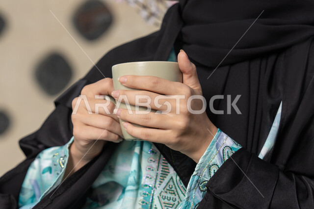 مطاعم  وكافيهات في الامارات العربية المتحدة، قضاء وقت الفراغ باحتساء المشروب المفضل, تناول الأعشاب الشتوية الدافئة، الاستمتاع بالمشروبات الساخنة، صورة مقربة لامراة اماراتية ترتدي العباءة والحجاب تجلس على اريكة مريحة تقوم بشرب القهوة في المقهى